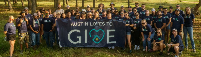 A large group of people all wearing a GIVE shirt, posing for a photo, holding up a banner that says Austin loves to GIVE