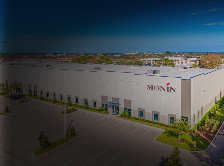 A large building with the Monin logo on the front, surrounded by a parking lot and trees