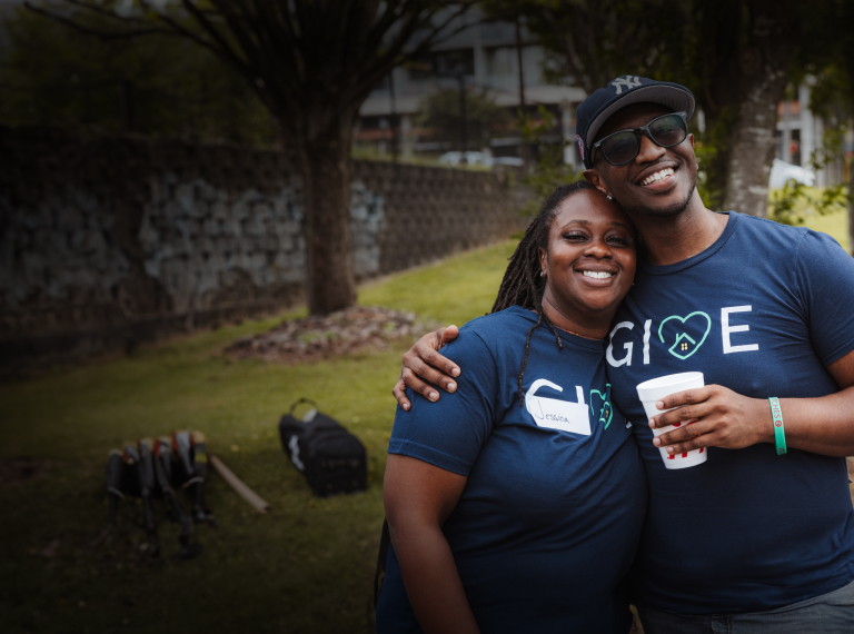 A man and woman wearing GIVE shirts, smiling and posing for a photo in front of trees