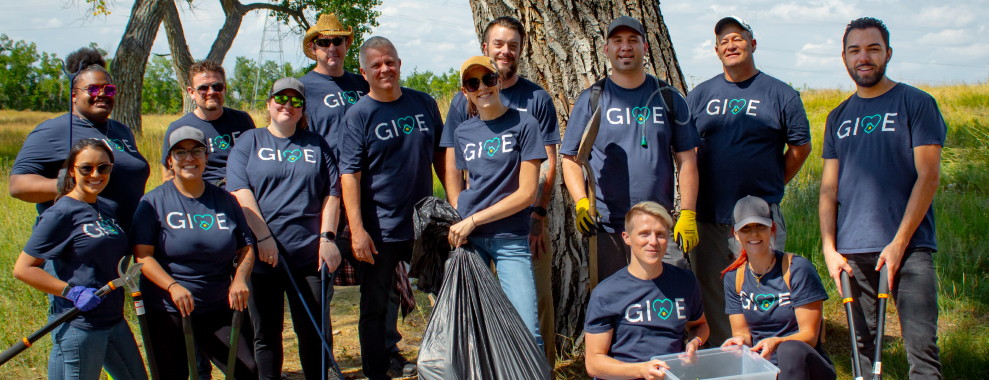 A varied group of people wearing GIVE shirts, smiling and posing for a photo. Many of them are holding gardening tools